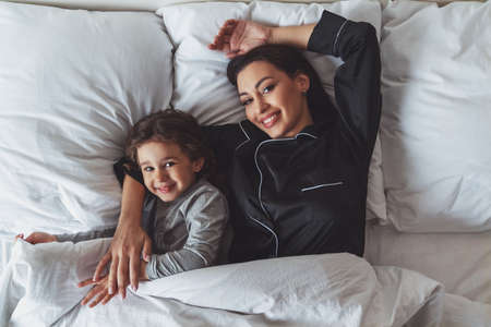 Happy morning. A young beautiful mother and her little daughter have just woken up in bed at home, a mother hugs her daughter with a smile.の写真素材