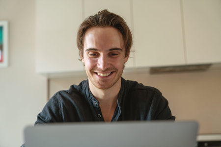 Young successful businessman at work sitting at kitchen at the table using laptop and drinking...