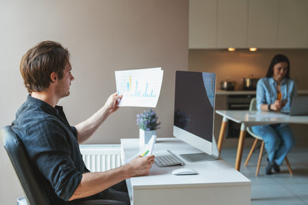 Young couple man and woman freelancers working using computer and laptop sitting in the kitchen at...