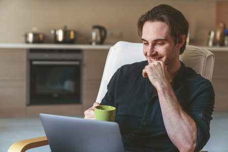 Young male businessman freelancer sitting in chair in kitchen at home using laptop and drinking tea...