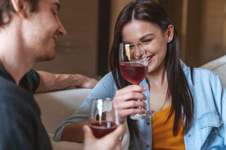 Young cheerful couple guy and girl sitting on sofa at home in apartment, holding glasses of wine and laughing looking at each other.の写真素材