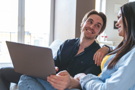 Young cheerful couple of guy and girl freelancers sitting on sofa at home in apartment using laptop and discussing something.の写真素材