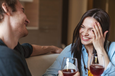 Young cheerful couple guy and girl sitting on sofa at home in apartment, holding glasses of wine and...