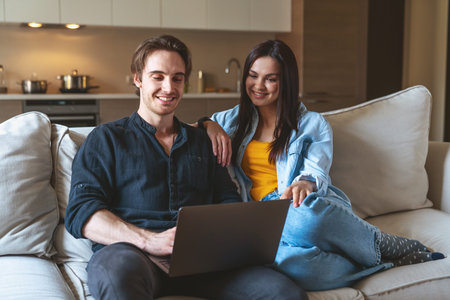 Young cheerful couple of guy and girl freelancers sitting on sofa at home in apartment using laptop...
