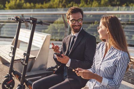 Young couple of tourists while talking sitting on a bench and drinking coffee to go while riding an electric scooter in the morning city.の写真素材
