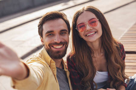Young couple having fun talking on a bench and taking a selfie using the phone while walking in the morning city.の写真素材
