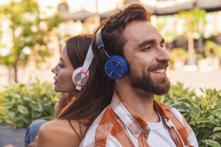 Young smiling couple listening to music on headphones sitting on a park bench back to back.