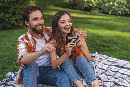 Young cheerful couple sitting on a blanket in the park during a picnic and drinking alcohol from a...