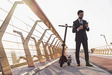 Young smiling bearded businessman in a business suit while riding an electric scooter on the background of the morning cityの写真素材