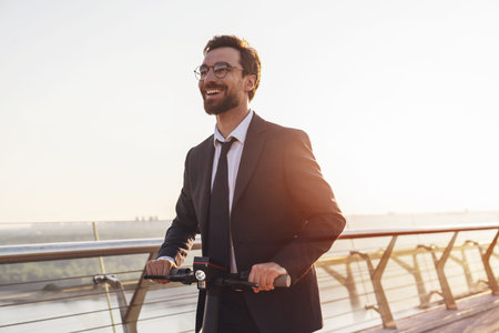 Young cheerful bearded businessman in a business suit while riding an electric scooter uses a...