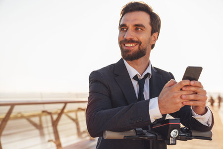 Young cheerful bearded businessman in a business suit while riding an electric scooter uses a smartphone on the background of the morning cityの写真素材