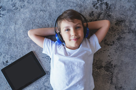 Portrait of a little boy listening to music on headphones and using an electronic tablet lying on...