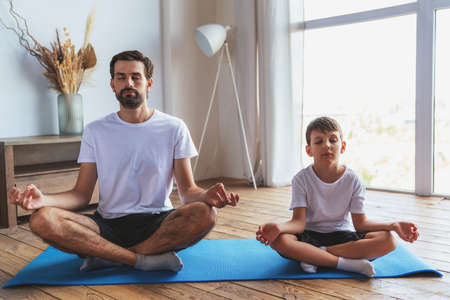 A young man helps his son during fitness training by doing yoga at home in the living room.の写真素材