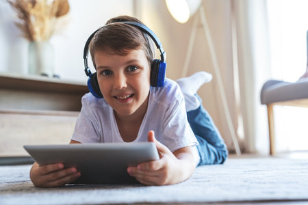 Portrait of a little boy listening to music on headphones and using an electronic tablet lying on...