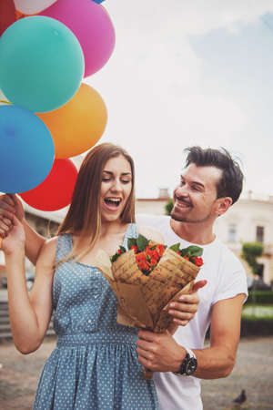 Romantic date outdoors. Handsome man surprised his woman with flowers and colorful balloons. Front view.の写真素材