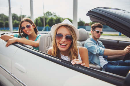 Three friends in cabriolet, shot outdoors. Attractive girls smiling at camera while theirs boyfriend driving the car.の写真素材