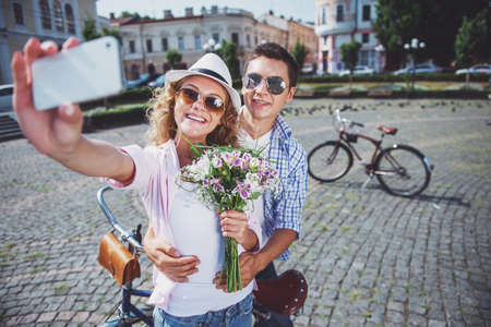 Young beautiful couple taking selfie. Man hugging woman with flowers in the city against bicycle on background.の写真素材