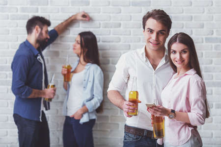 Smiling young couple holding a bottle of beer and a smartphone, the other a young couple with a bottle of beer in the background on a brick wall backgroundの写真素材