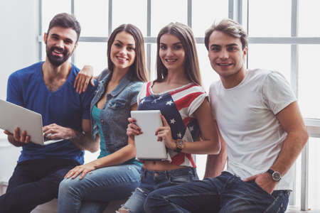Four young attractive people using laptop and tablet PC, sitting on the window-sill, smilingの写真素材