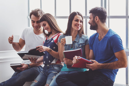 Four young attractive people sitting on the window sill, smiling, keeping in hands cup and notebook