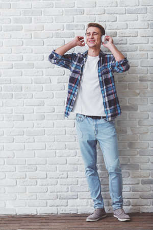 Young handsome man dressed in fashionable clothes, listening to music with headphones on mobile phone, against a brick wallの写真素材