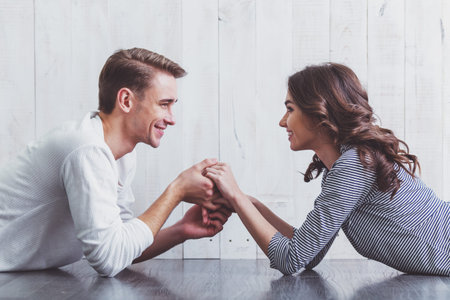Young beautiful couple lying on Wooden floor, holding hands, looking at each other, smiling at home