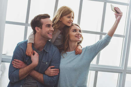 Beautiful young family smiling and making selfie while sitting near the window at homeの写真素材
