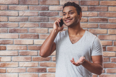 Young handsome Afro-American man talking on the phone and smiling while standing against brick wallの写真素材