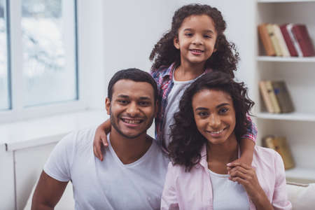 Cute little Afro-American girl and her beautiful young parents hugging, looking at camera and smiling while sitting on a sofa in the room.の写真素材