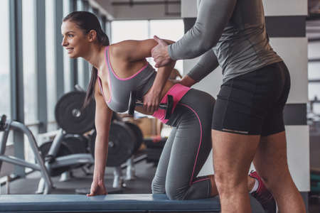 Attractive young woman working out with dumbbells in gym, handsome muscular trainer helping herの写真素材