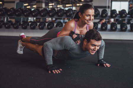 Attractive young muscular man doing push ups in gym, woman lying on him. Both smiling