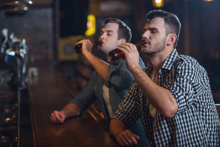 Two young men in casual clothes are drinking beer while sitting at bar counter in pubの写真素材