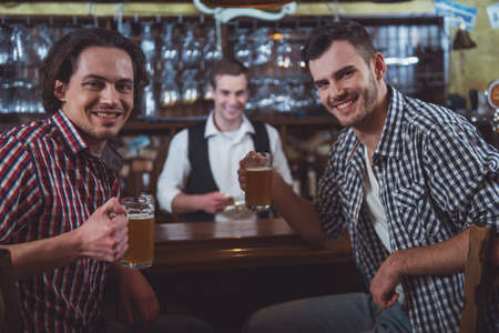 Two men are smiling, looking at camera and holding glasses of beer while sitting at bar counter in pub, a bartender in the backgroundの写真素材