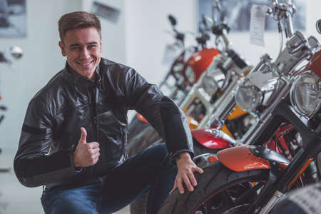 Attractive young blond man in black leather jacket is looking at camera, showing Ok sign and smiling...