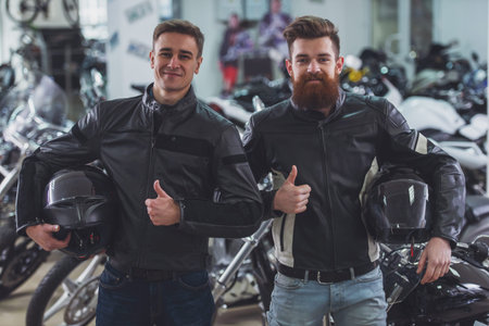 Handsome young bearded and blond men in black leather jackets are holding helmets, showing Ok sign...