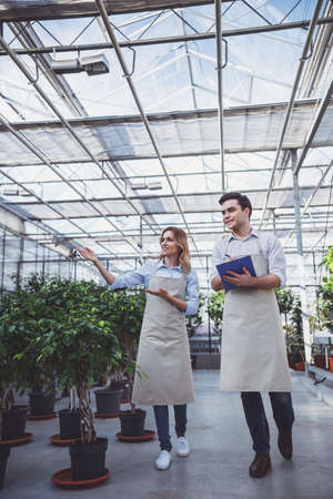 Beautiful young woman and man in aprons are making notes and smiling while examining plants in orangeryの写真素材
