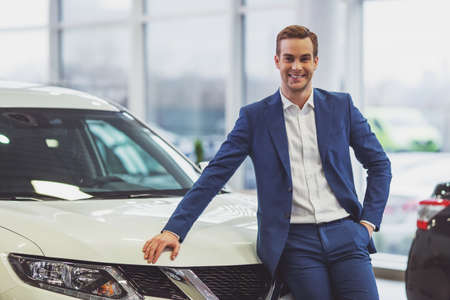 Handsome young businessman in classic blue suit is smiling and looking at camera while leaning on a car in a motor showの写真素材
