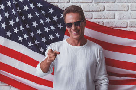 Handsome man in white sweatshirt and sunglasses is smiling, pointing and looking at camera while standing against American flagの写真素材