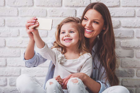 Beautiful young mother is making a selfie with her cute little daughter using a smart phone and smiling, against white brick wallの写真素材