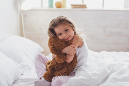 Sweet little girl is hugging a teddy bear, looking at camera and smiling while sitting on her bed at homeの写真素材