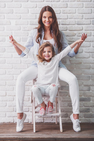 Beautiful young mother and her cute little daughter are looking at camera and smiling, sitting against white brick wallの写真素材