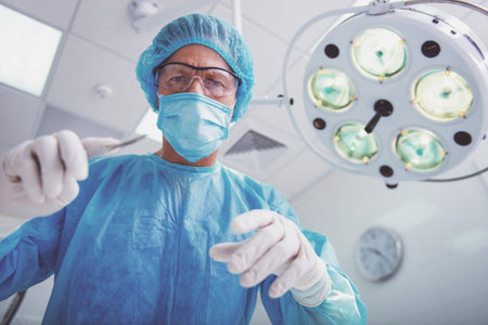 Surgeon in surgical clothes is holding a scalpel ready to operate and looking at camera, standing in...