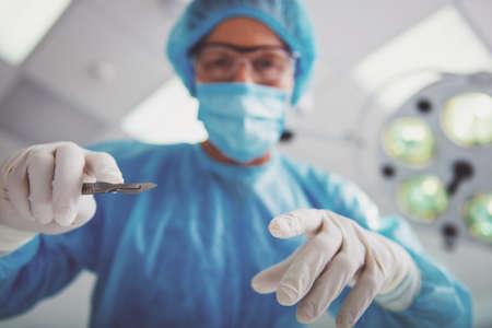 Surgeon in surgical clothes is holding a scalpel ready to operate and looking at camera, standing in a modern operating room, bottom viewの写真素材