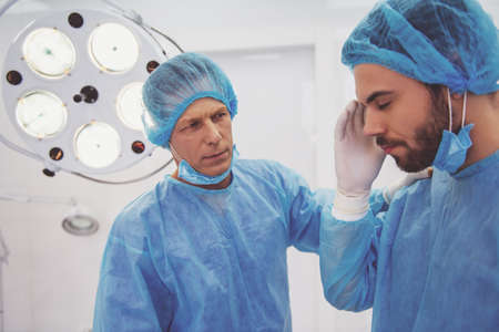 Handsome young doctor is having a headache, his colleague is calming him, standing in a modern operating roomの写真素材
