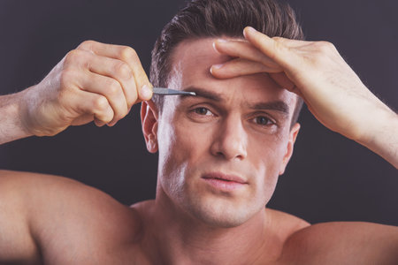 Portrait of handsome naked man plucking his eyebrows, on a dark backgroundの写真素材