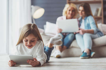 Little girl is using a tablet, in the background her mom and granny are using a laptop while sitting on sofa at homeの写真素材