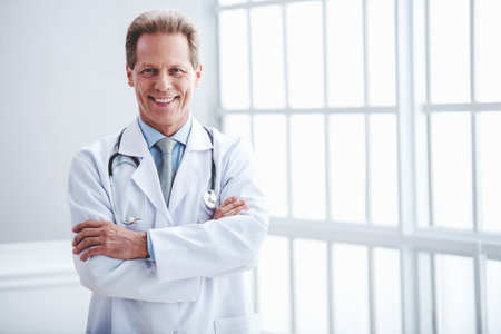 Handsome middle aged doctor in white medical coat is looking at camera and smiling while standing with crossed armsの写真素材