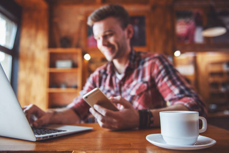 Cup of coffee on bar counter, in the background young businessman is using a laptop and smiling, sitting in a modern urban cafeの写真素材