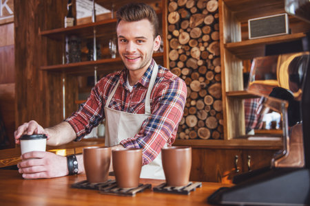 Handsome young barista in apron is covering a cup of coffee, looking at camera and smiling while standing at the bar counter at cafeの写真素材