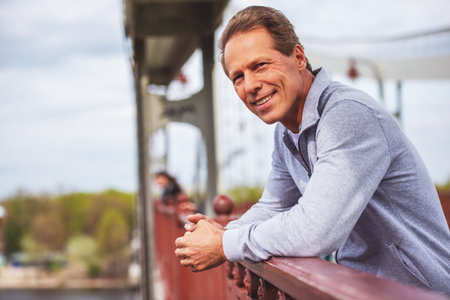 Handsome middle aged man in sports uniform is leaning on bridge, looking at camera and smiling while...
